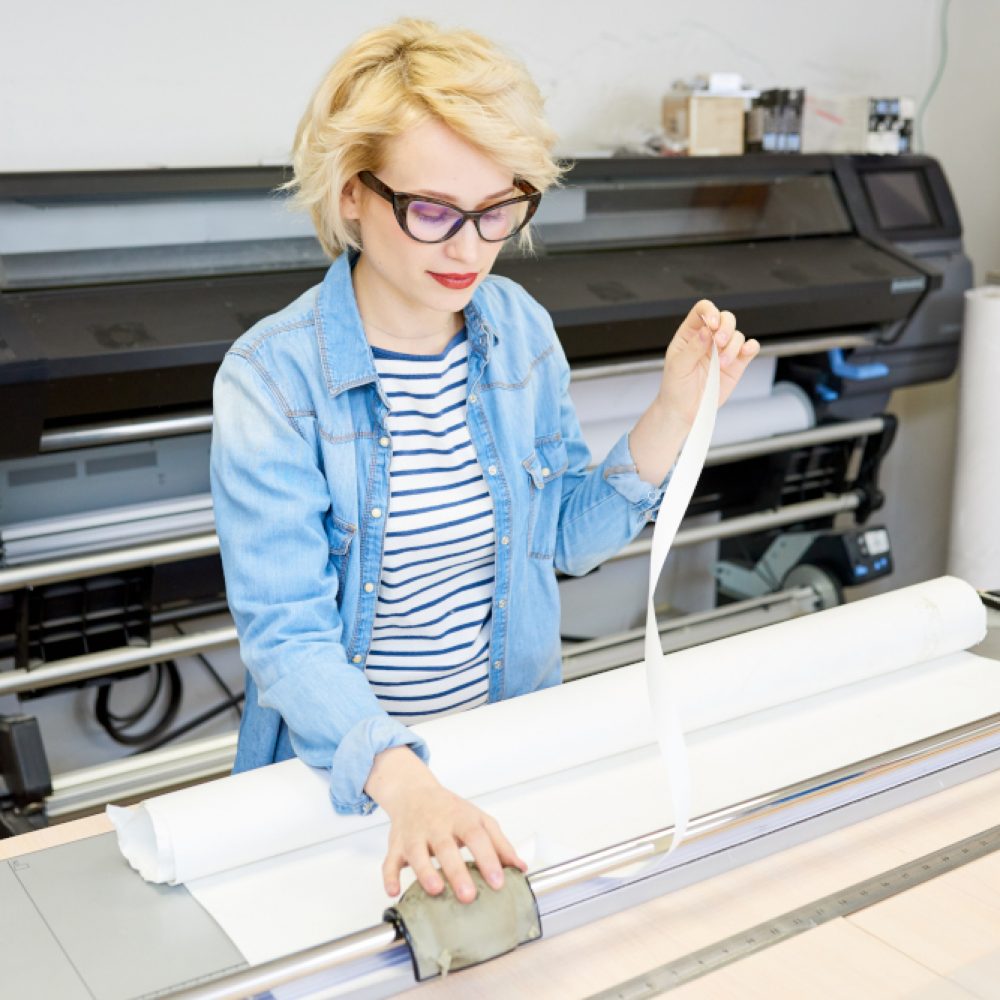 Beautiful young woman in glasses printing drafts on plotter during work in office.